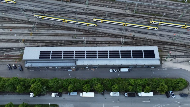 Luftaufnahme: Solaranlage auf dem Dach einer langen Halle neben Bahngleisen